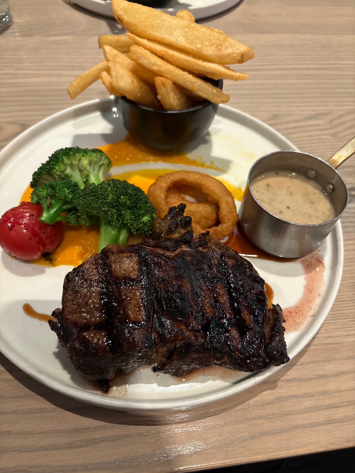 A steak dinner with chips, broccoli, roasted tomato, an onion ring, and peppercorn sauce, photographed from above